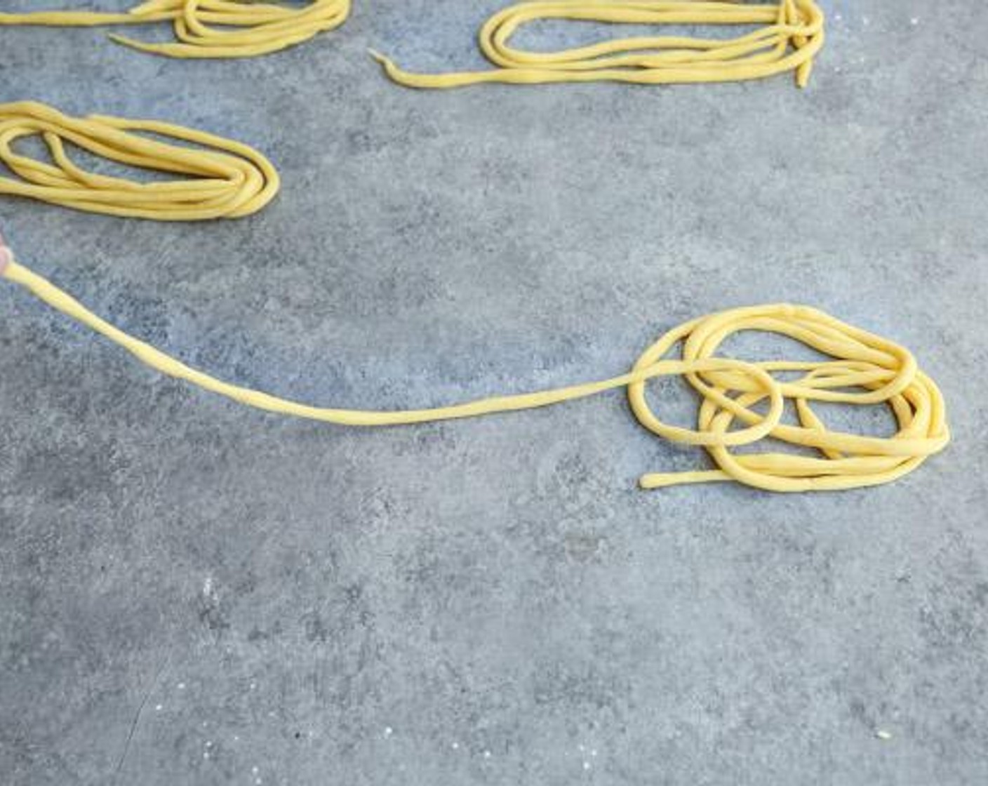 Schritt 6 Am Ende soll der fertige Strang wie eine Halskette 5 Mal um den Hals gelegt werden können. Die gewickelte Nudel auf ein Blech oder ein Brett geben und mit einem Küchentuch abdecken, bis man den Vorgang mit den restlichen Teigstücken wiederholt hat.