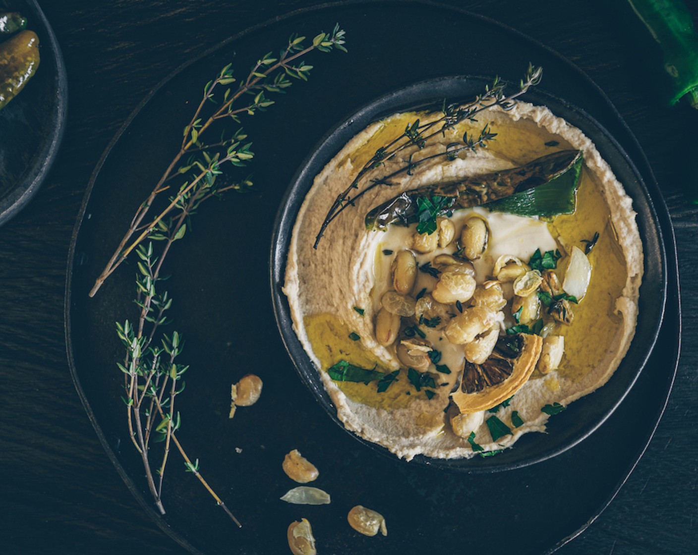 Schritt 6 Anrichten: Den Hummus in einen Teller geben, mit einem Löffel eine kleine Mulde formen und Tahini (wenig) hineingeben. Danach mit den angebratenen Bohnen, Jalapeños, der Salzzitrone (1/2 Scheibe) und der Petersilie (wenig) garnieren. Zum Schluss Olivenöl (nach Bedarf) darübergeben und servieren.