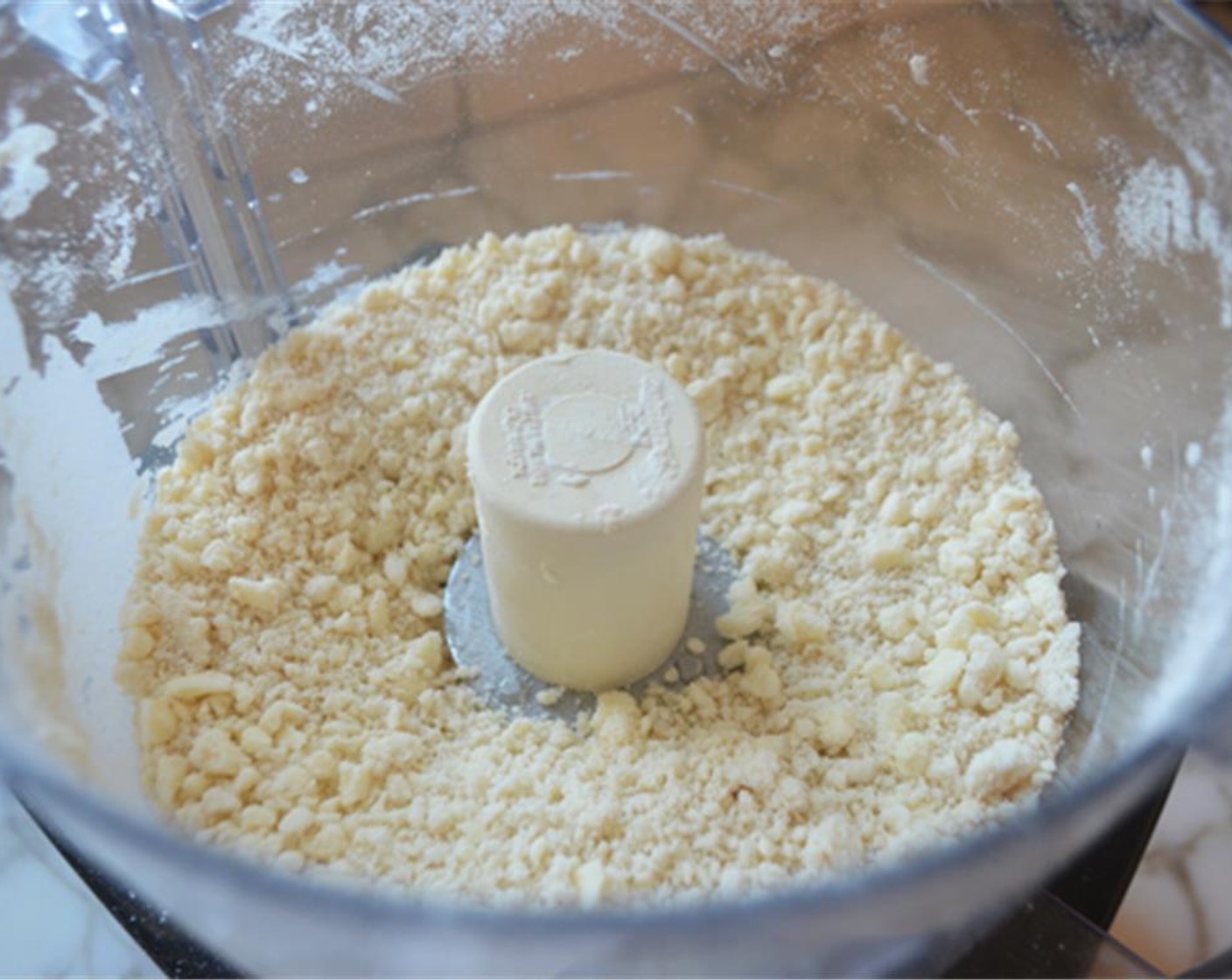 Schritt 3 Die Hälfte des Wassers (4 Esslöffel) hinzufügen und einige Male pulsieren lassen, bis es eingearbeitet ist. Das restliche Wasser hinzufügen und pulsieren, bis die Mischung gleichmässig angefeuchtet und sehr krümelig ist.