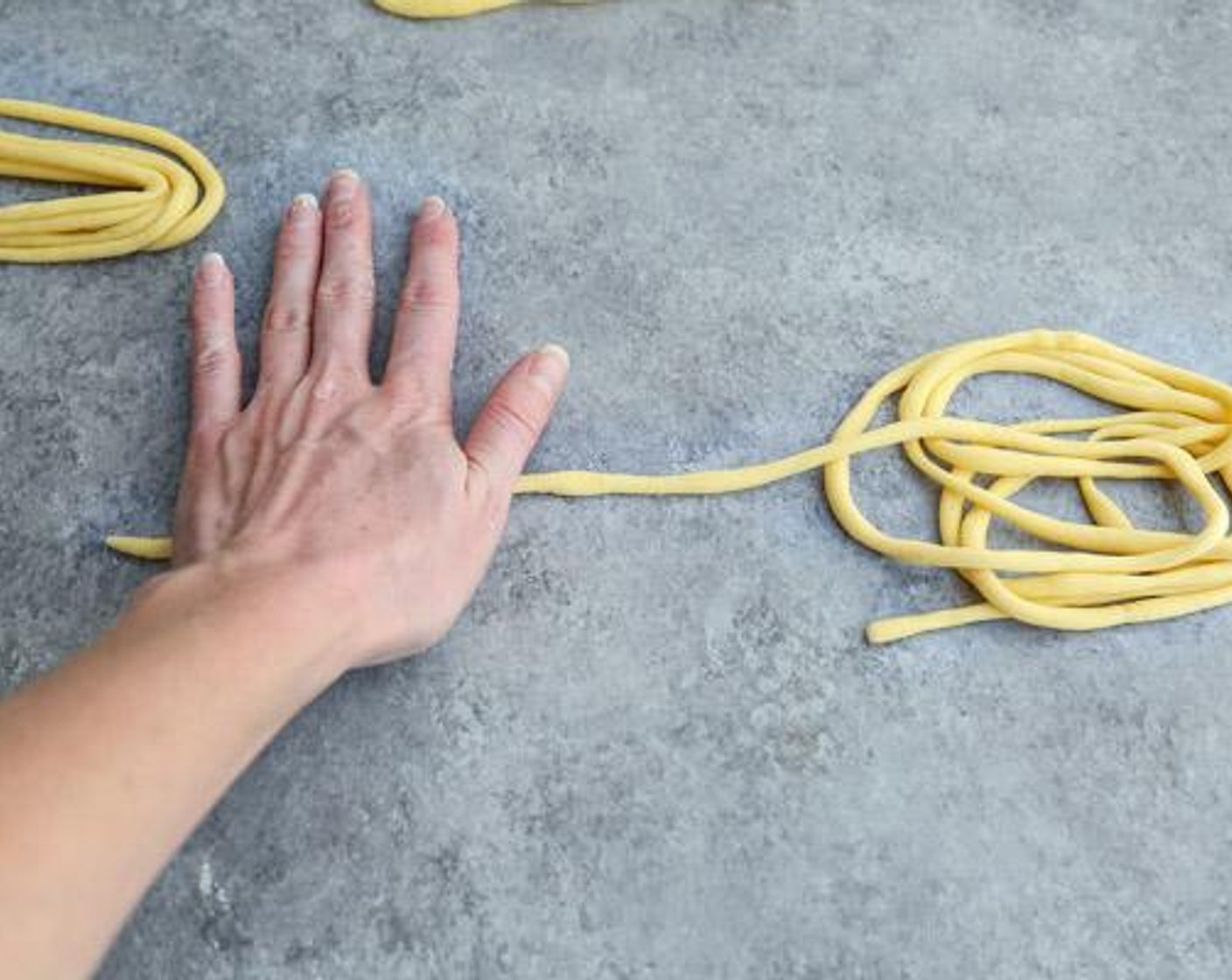 Schritt 5 Während man mit der rechten Hand rollt und dabei ausreichend Druck auf den Teig ausübt, wird mit der linken Hand, Reibung erzeugt um den Strang zu verlängern. Langsam durch den Teig arbeiten und den fertigen, dünnen Teil des Strangs in Windungen nach rechts drapieren. Falls ein Stück reisst, diesen einfach wieder befestigen weiter rollen.