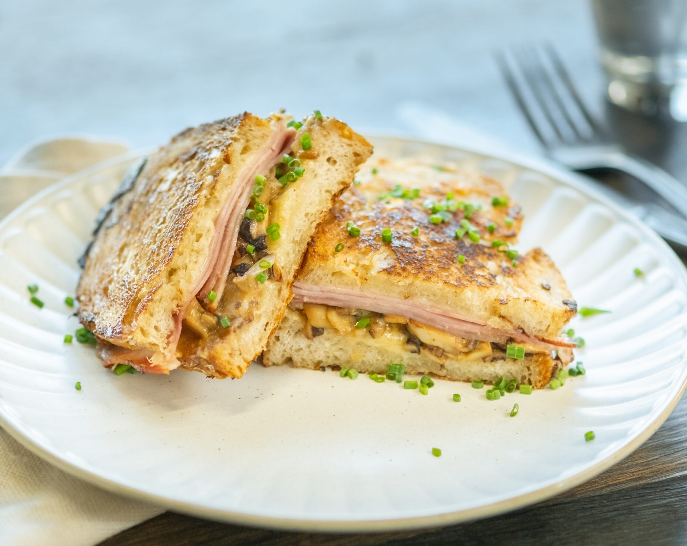 Schritt 3 Sandwiches: Sauerteigbrot (8 Scheiben) auf einer Seite mit Butter (2 2/3 Esslöffel) bestreichen. 4 Scheiben mit Butter nach unten auf die Arbeitsfläche legen, mit der Hälfte des geriebenen Gruyère (200 g) bestreuen, mit je 2 Scheiben Kochschinken (8 Scheiben) belegen. Grosszügig Pilzragout darauf verteilen. Erneut Käse darüberstreuen, etwas Käse übrig lassen. Mit den restlichen Brotscheiben bedecken, gebutterte Seite nach aussen.